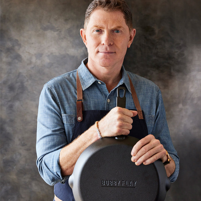 A man in a denim shirt and apron holds the Bobby Flay by GreenPan 10" Cast Iron Frypan, standing before a gray textured background.
