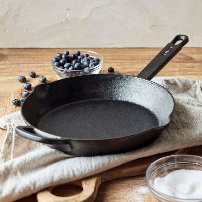 The Bobby Flay by GreenPan 10" Cast Iron Frypan, featuring a black finish and roomy cooking surface, rests on a beige cloth with a small bowl of blueberries and sugar nearby on wood. Blueberries are scattered around the pan.