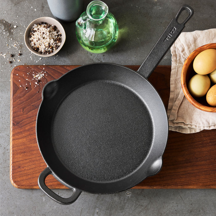 The Bobby Flay by GreenPan 10" Cast Iron Frypan in black rests on a wooden cutting board, near a bowl of eggs, oil, and coarse salt with pepper atop a gray kitchen countertop.