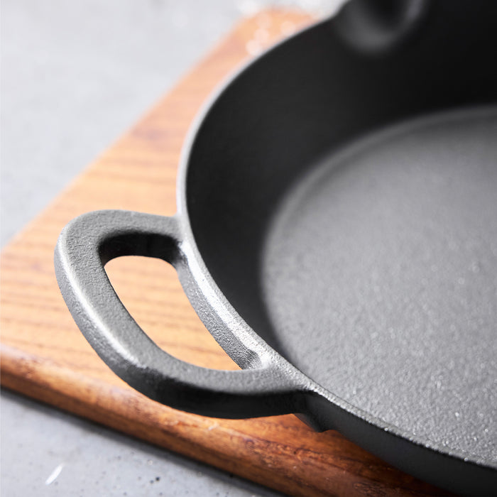 Close-up of the Bobby Flay by GreenPan 10" Cast Iron Frypan, featuring a black textured surface and handle, resting on a wooden cutting board—an heirloom-quality skillet perfect for any kitchen.