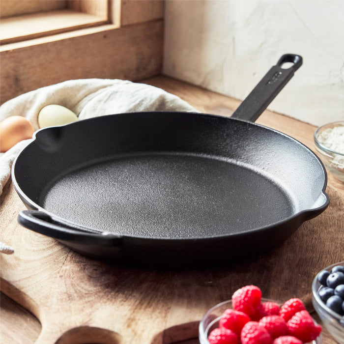 The Bobby Flay by GreenPan 12" Cast Iron Frypan, in black, sits on a wooden countertop near a window, surrounded by eggs, berries, oats in a bowl, and a light-colored cloth.