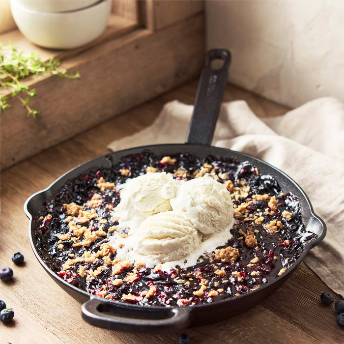 A Bobby Flay by GreenPan 12" Cast Iron Frypan holds a blueberry crumble topped with three vanilla ice cream scoops, displayed on a wooden surface with fresh blueberries scattered around.