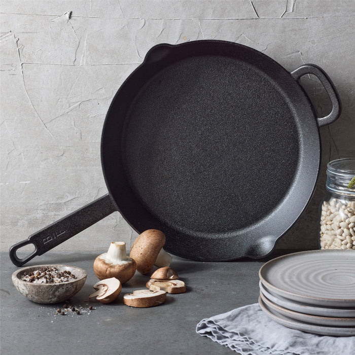 A Bobby Flay by GreenPan 12" Cast Iron Frypan in black leans against a textured gray wall, with sliced mushrooms, a bowl of pepper, stacked gray plates, a cloth, and a jar of white beans arranged on the gray surface in front.