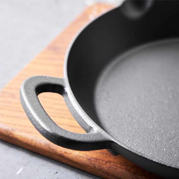 Close-up of the Bobby Flay by GreenPan 12" Cast Iron Frypan with a handle, resting on a wooden cutting board atop a gray surface.