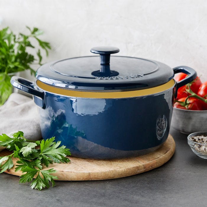 Blue Dutch oven with lid on a wooden board, surrounded by parsley, tomatoes, and assorted spices.