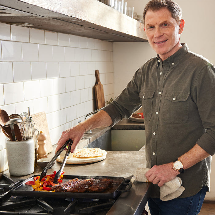 A man in a green shirt smiles while cooking steak and veggies in a pan on a stovetop, using the Bobby Flay by GreenPan 9.5" x 16.5" Reversible Cast Iron Grill Griddle. He holds tongs and a towel in his hands in a bright kitchen.