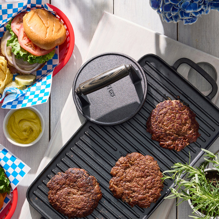 Three cooked burger patties on a grill pan with the Bobby Flay by GreenPan 6.5" Cast Iron Round Grill Press, next to baskets of buns, lettuce, tomato, onion, mustard, blue flowers, and rosemary on a white table.