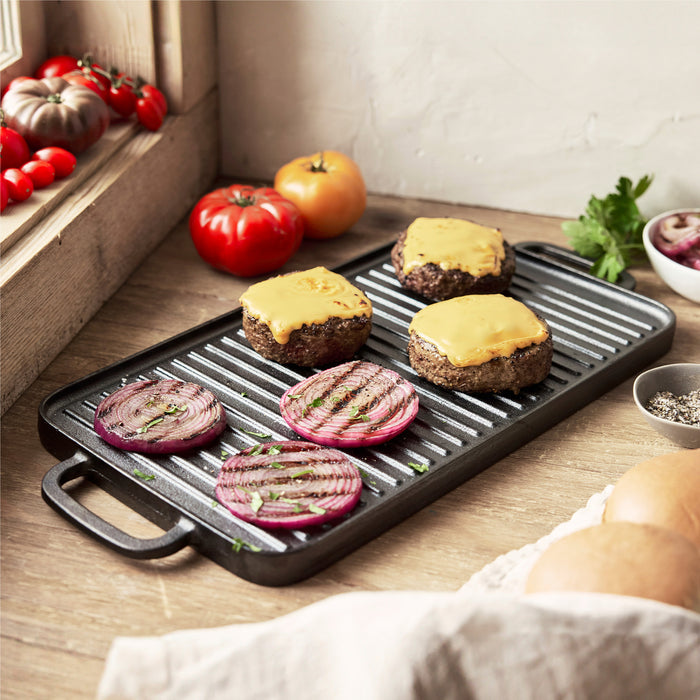 The Bobby Flay by GreenPan 9.5" x 16.5" Reversible Cast Iron Grill Griddle holds cheeseburgers and grilled red onion slices, surrounded by fresh tomatoes, parsley, buns, and a bowl of sliced red onions on a wooden counter.