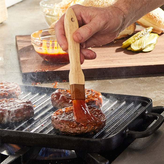 A wooden brush spreads sauce on burger patties cooking on the Bobby Flay by GreenPan 9.5" x 16.5" Reversible Cast Iron Grill Griddle, with pickles, buns, and sauce nearby—ideal for family-sized meals.