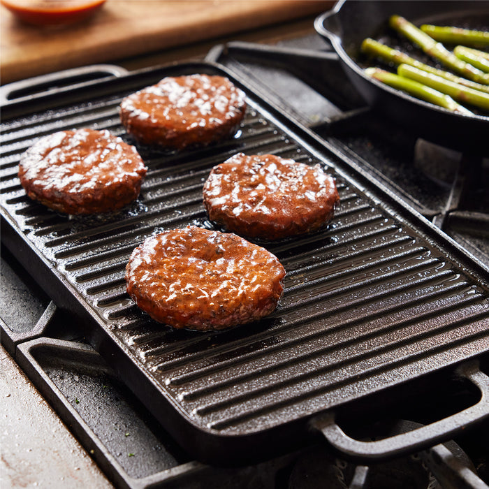 Four burger patties sizzle on the Bobby Flay by GreenPan 9.5" x 16.5" Reversible Cast Iron Grill Griddle, ideal for family meals, while a skillet cooks asparagus and a wooden board displays a small bowl of sauce nearby.