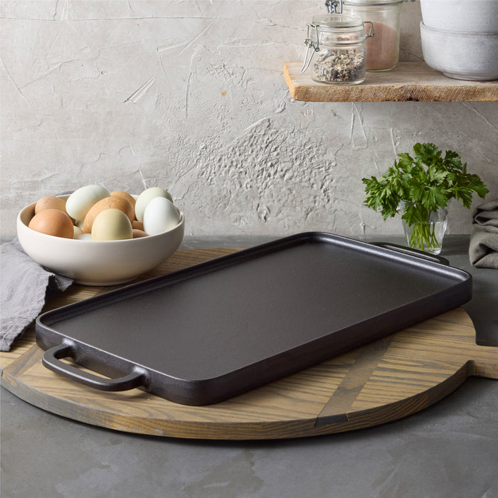 The Bobby Flay by GreenPan 9.5" x 16.5" Reversible Cast Iron Grill Griddle rests on a round wooden board, with assorted eggs, parsley, a gray cloth, and ingredient jars nearby—ideal for family-sized meals.