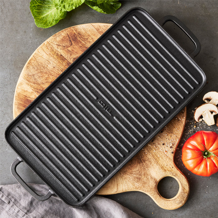 The Bobby Flay by GreenPan 9.5" x 16.5" Reversible Cast Iron Grill Griddle rests on a round wooden cutting board, surrounded by a tomato, mushrooms, lettuce, and a gray cloth—ideal for effortless family-sized meals.