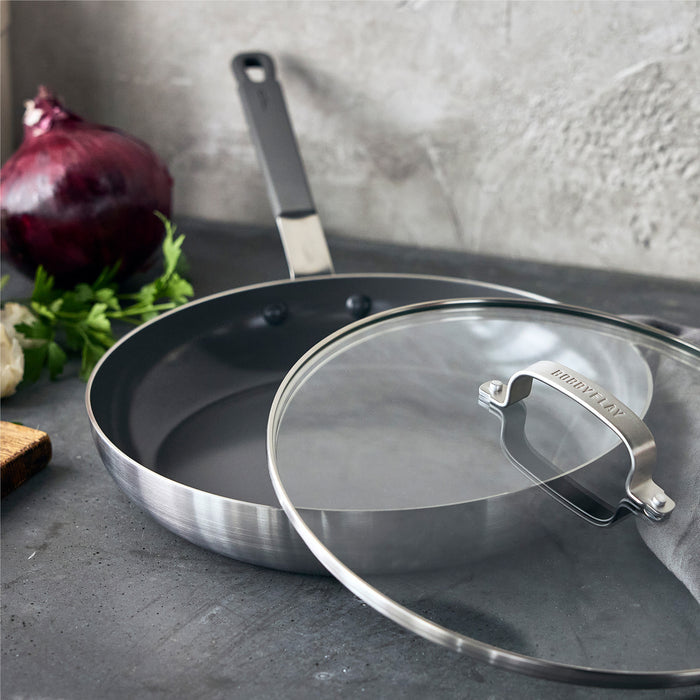 GreenPan 12-inch nonstick frypan with glass lid on a counter next to an onion and fresh herbs.