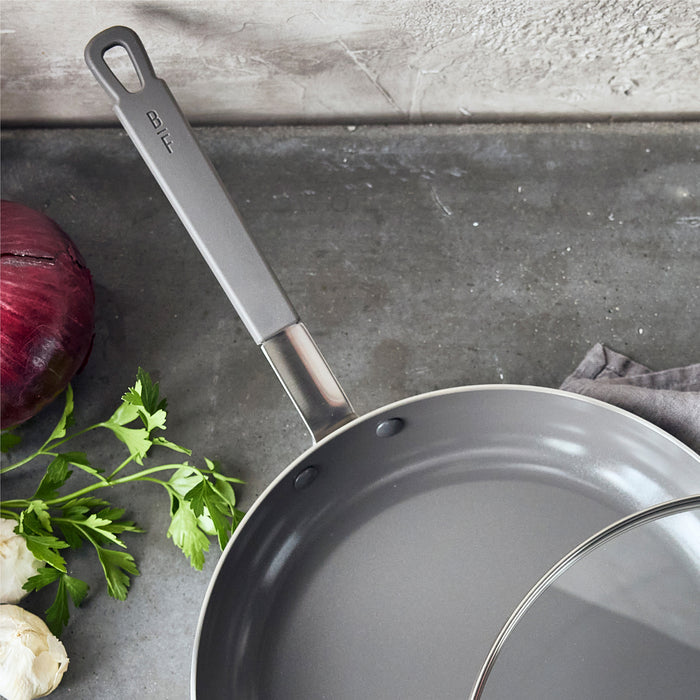 12-inch nonstick frypan with lid on a kitchen counter, shown with parsley, garlic, and red onion nearby.
