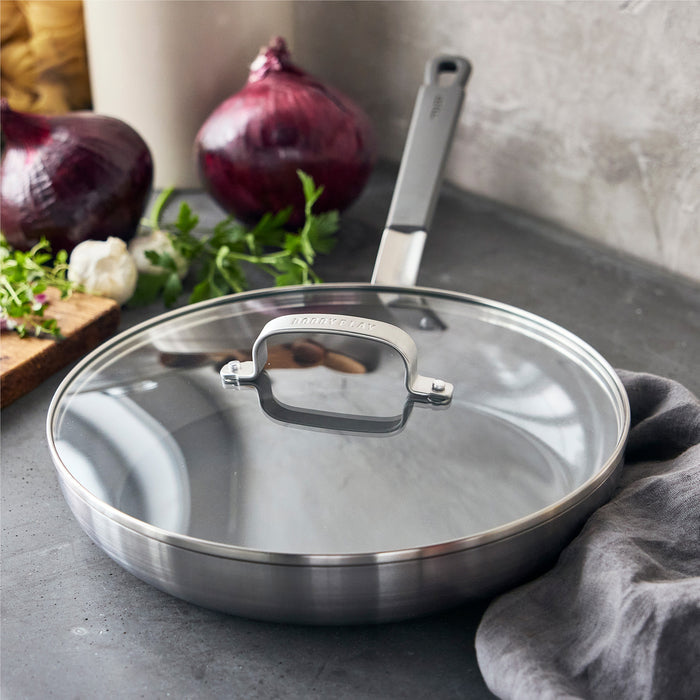 Gray nonstick frypan with lid on a counter, shown next to onions, herbs, and a gray cloth.