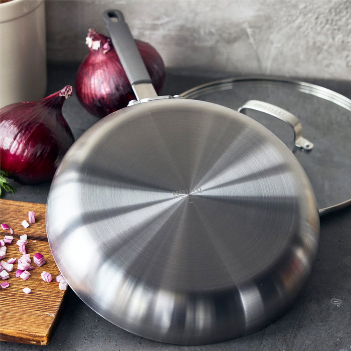 Stainless steel frypan with lid on a counter, next to whole and chopped red onions.