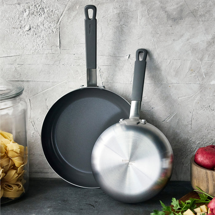 Stainless steel 8-inch and 10-inch frypan set on a counter with pasta, fresh herbs, and potatoes.