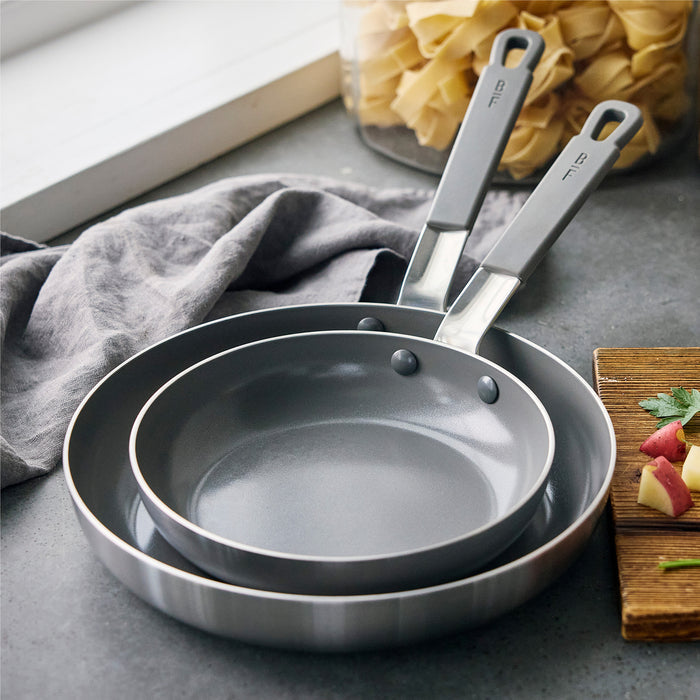 Two gray GreenPan frypans, 8-inch and 10-inch, stacked on a kitchen counter with a gray towel nearby.