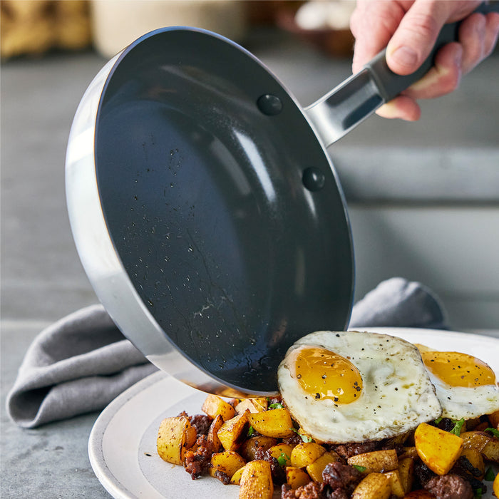 Hand holding a stainless steel nonstick frypan above a plate with eggs and assorted vegetables.