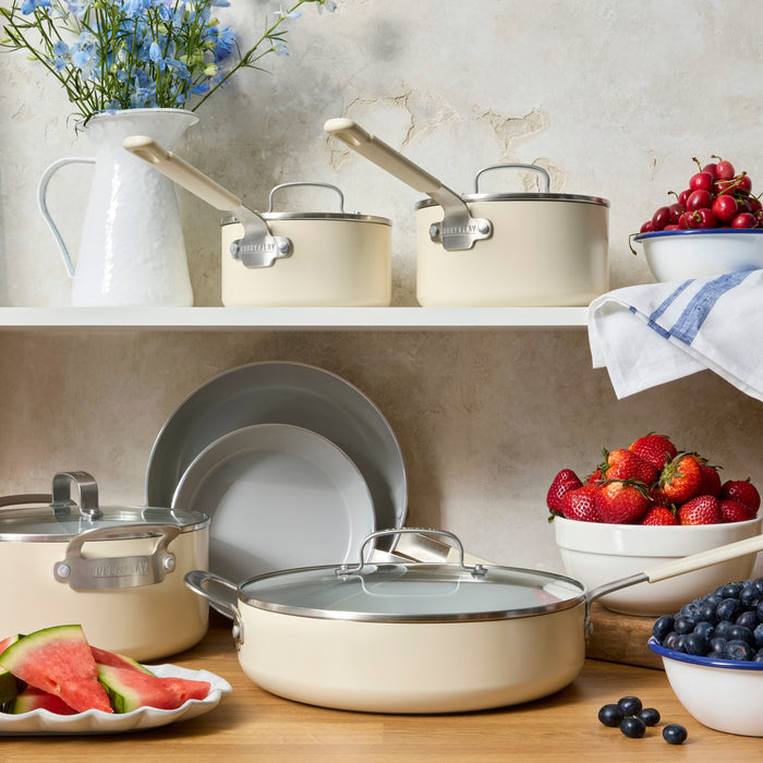 Cream-colored sauté pan on a kitchen shelf next to fruit bowls, a white pitcher, and stacked plates.
