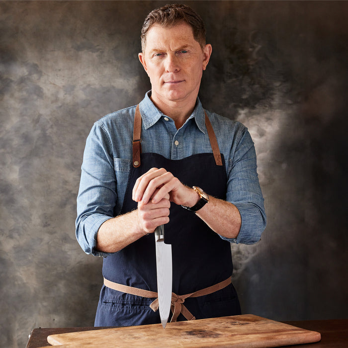 A man in a denim shirt and apron stands behind a wooden cutting board, gripping a knife from the Bobby Flay by GreenPan Stainless Steel 13-Piece Knife Block Set, blade pointed down, against a dark textured background.