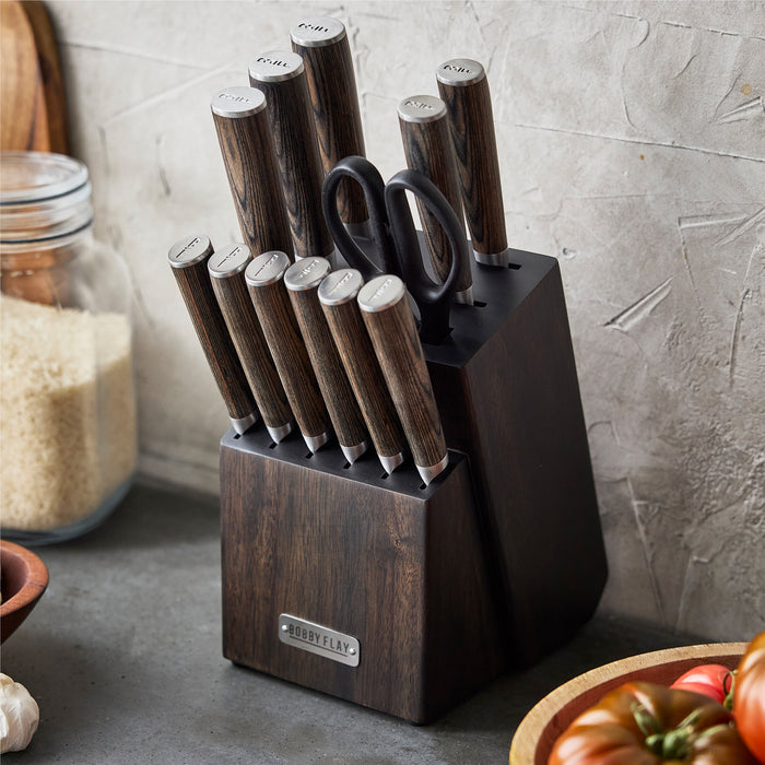 A Bobby Flay by GreenPan Stainless Steel 13-Piece Knife Block Set with wooden handles and stainless steel blades sits on a kitchen counter beside a jar of rice and a bowl of vegetables.