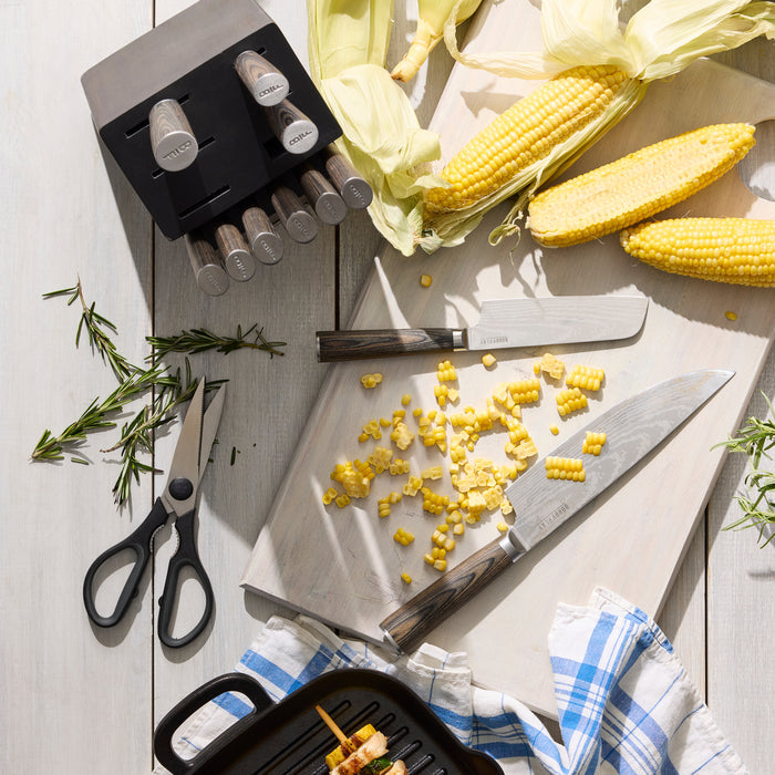 A kitchen scene with the Bobby Flay by GreenPan Stainless Steel 13-Piece Knife Block Set, featuring knives, scissors, a knife block, corn on the cob and kernels on a cutting board, rosemary sprigs, and a grill pan on a light wooden surface.