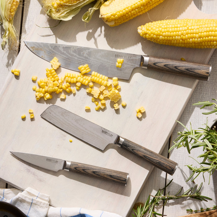 Three knives from the Bobby Flay by GreenPan Stainless Steel 13-Piece Knife Block Set rest on a cutting board with fresh corn cobs, herbs, and some chopped corn kernels scattered around.