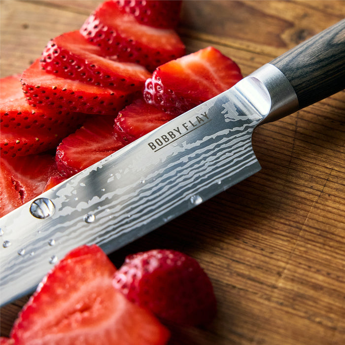 A close-up of a chef’s knife from the Bobby Flay by GreenPan Stainless Steel 13-Piece Knife Block Set, featuring a textured blade engraved with BOBBY FLAY, slices fresh strawberries on a wooden cutting board.