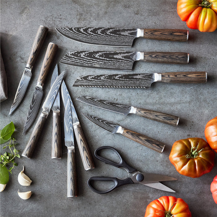 The Bobby Flay by GreenPan Stainless Steel 13-Piece Knife Block Set features Damascus-patterned blades and wooden handles, shown with kitchen shears, garlic, herbs, and heirloom tomatoes on a gray countertop.