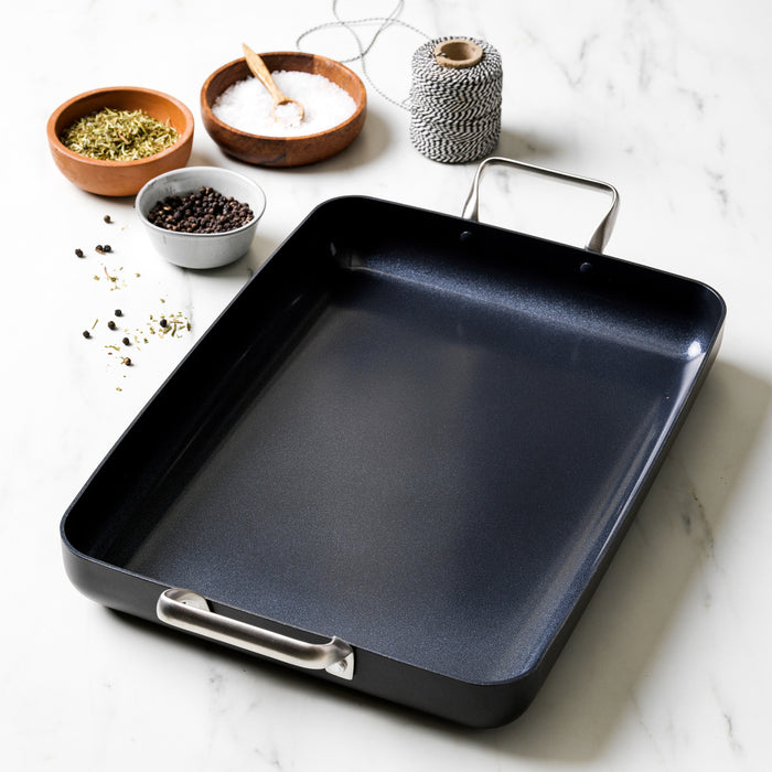 Black ceramic nonstick 13.8″ x 10″ rectangular roasting pan with metal handles on a marble surface with spices and twine.