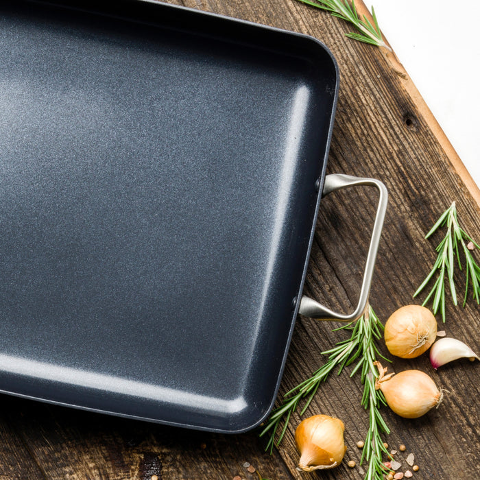 Black aluminum 13.8″ x 10″ ceramic nonstick roasting pan on a wood surface with rosemary, shallots, and garlic.