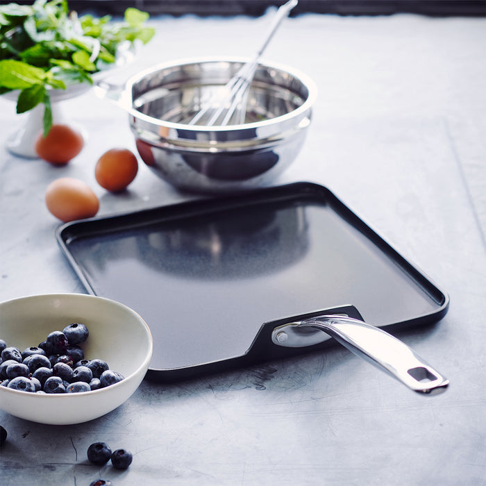 Black ceramic square griddle pan with metal handle, next to blueberries, eggs, bowls, and a whisk.