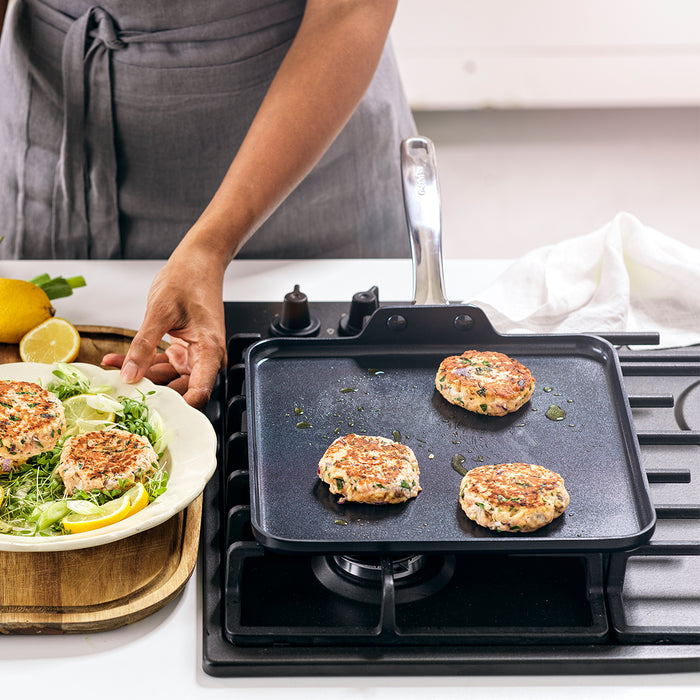 Black ceramic square griddle with a salmon patty, fresh greens, and lime wedges on top.