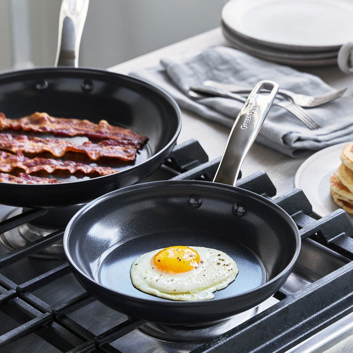 Fried egg and bacon cooking in a black ceramic 8-inch frypan on a stove, with plates next to the pan.