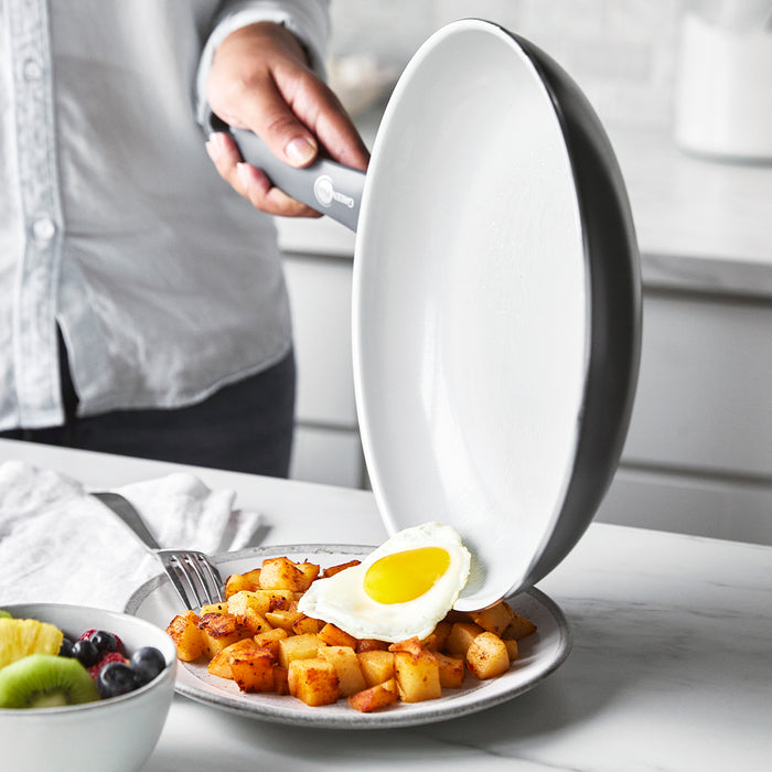 Fried egg being slid from charcoal gray ceramic nonstick frypan onto potatoes, with fruit nearby.