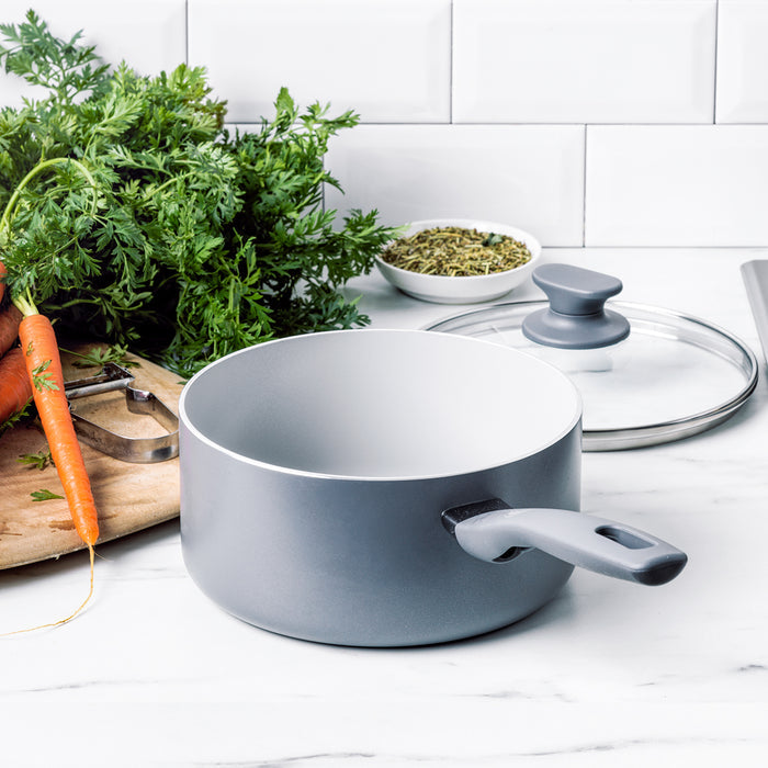 Gray ceramic nonstick saucepan with lid on a kitchen counter next to vegetables and a wooden cutting board.