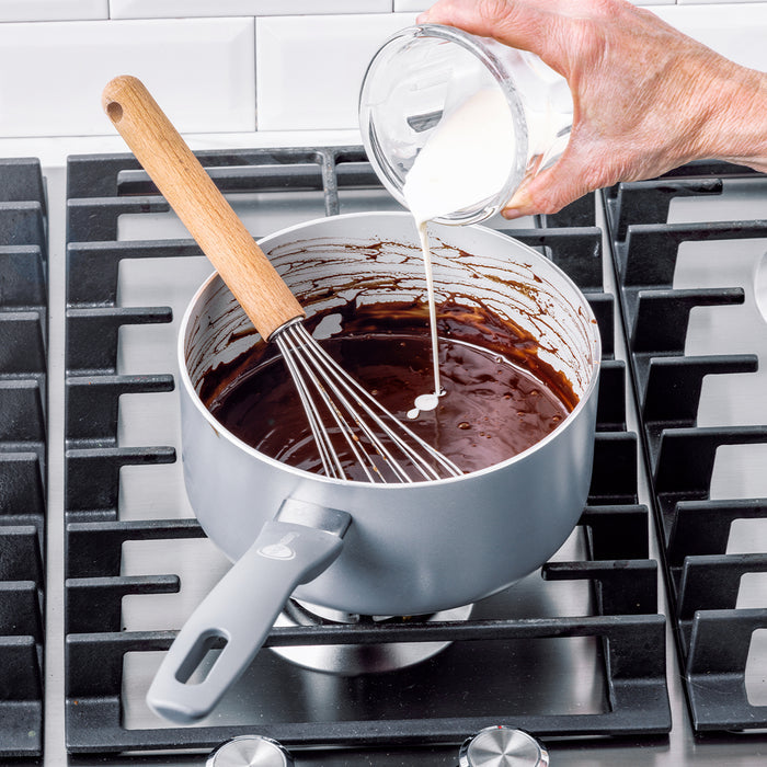 Cream being poured into melted chocolate in a black Dover ceramic saucepan, with a metal whisk resting inside.