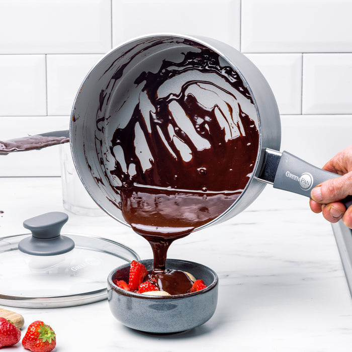 Chocolate sauce being poured from a Dover ceramic nonstick saucepan onto strawberries in a bowl on a white kitchen counter.
