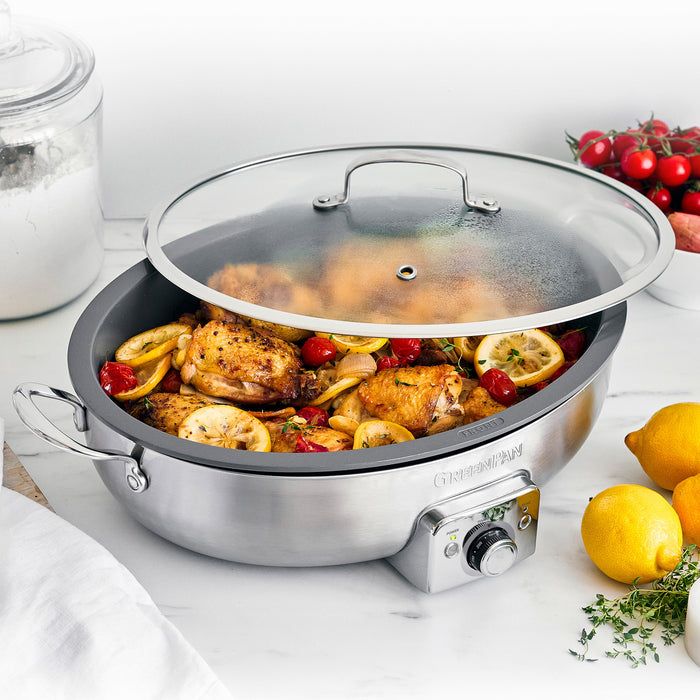 Oval electric skillet with glass lid cooking chicken, tomatoes, and lemons on a kitchen countertop.
