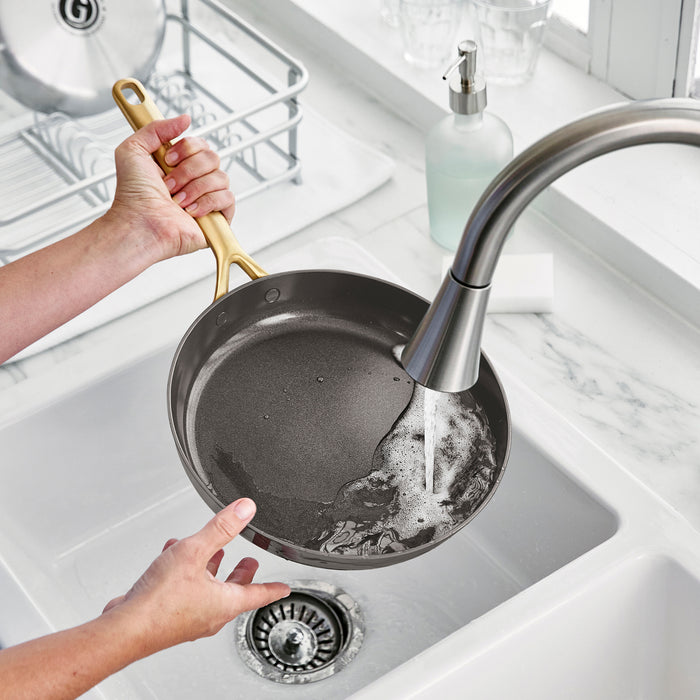 Hand rinsing a set of cocoa-colored ceramic nonstick frypans with champagne handles under a kitchen faucet.