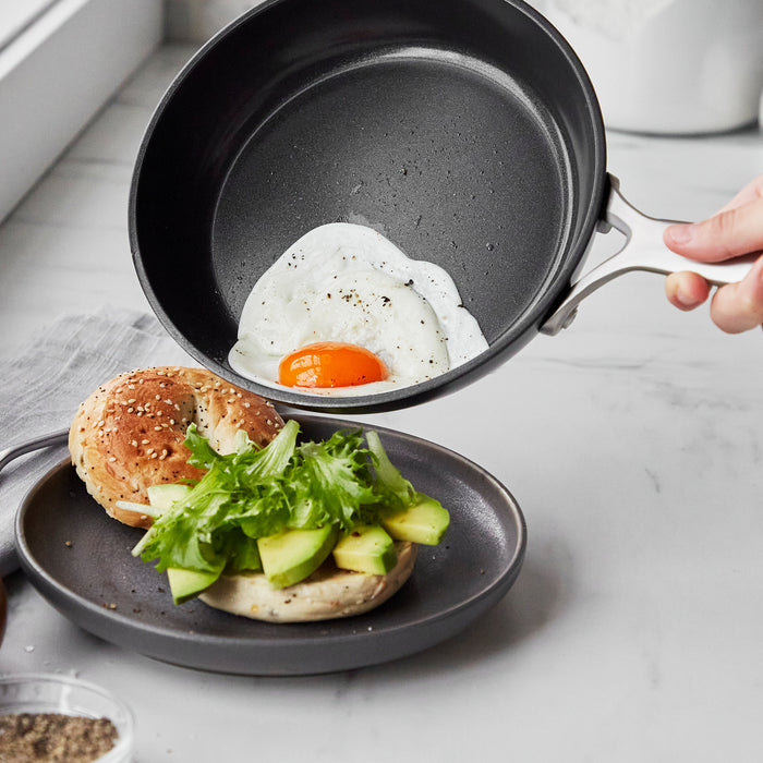 A fried egg slides from a white ceramic nonstick frypan onto a bagel topped with avocado and greens.