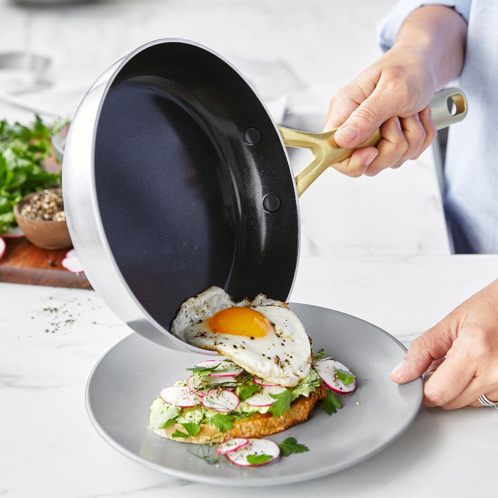 A sunny-side up egg slides from a stainless steel frypan with champagne handles onto avocado toast.