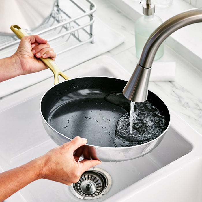 Stainless steel frypan with champagne handle being rinsed under a kitchen faucet in a white sink, soap bar nearby.