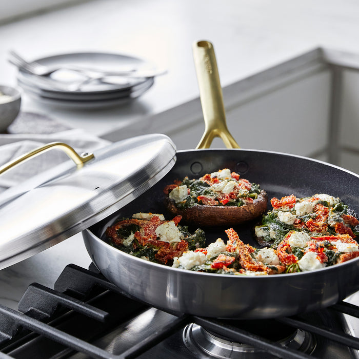 Stainless steel frypan with lid and champagne handles on stove, filled with stuffed mushrooms, greens, cheese, and peppers.