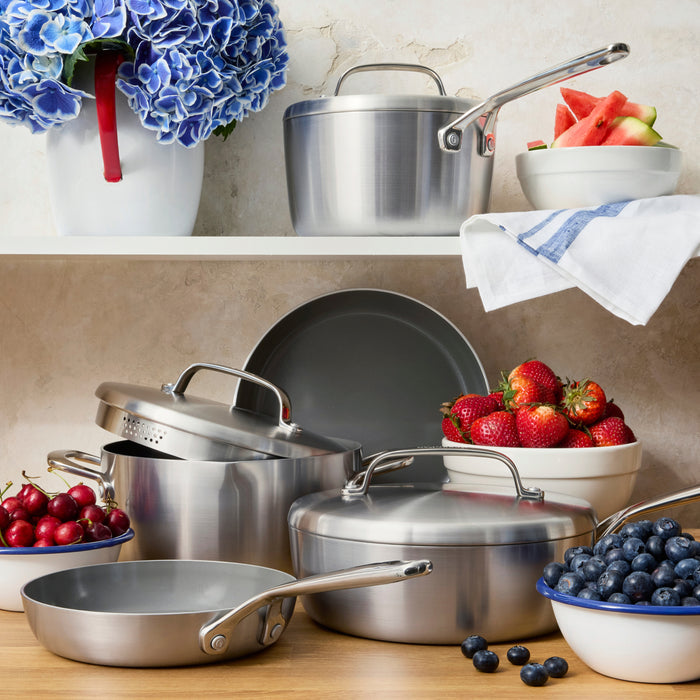 Stainless steel 10-piece cookware set with mirror handles on a counter, with fruit bowls and blue flowers in the background.
