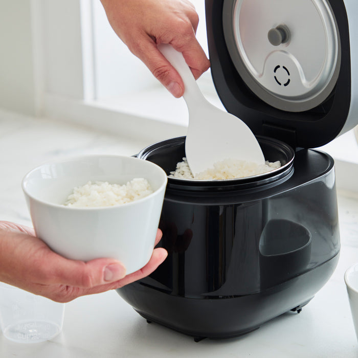 Black GreenLife 4-cup rice and grains cooker with rice being scooped into a white bowl using a white spatula.