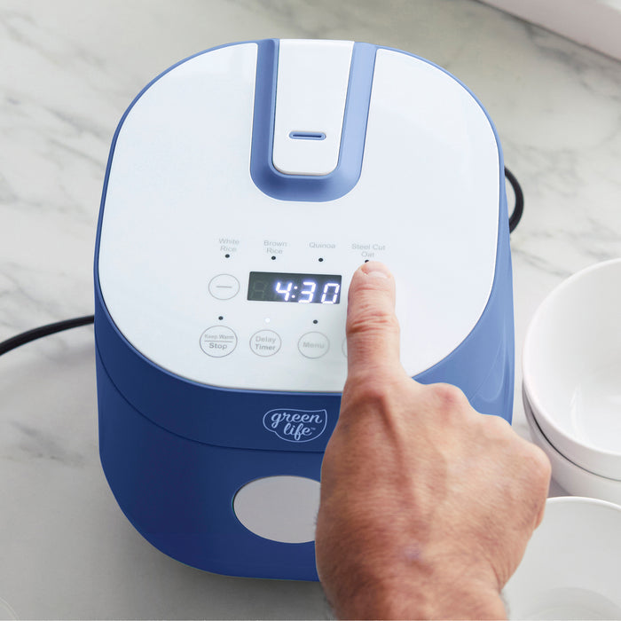 A hand presses a button on a blue 4-cup GreenLife rice and grains cooker showing 4:30 on a kitchen counter.
