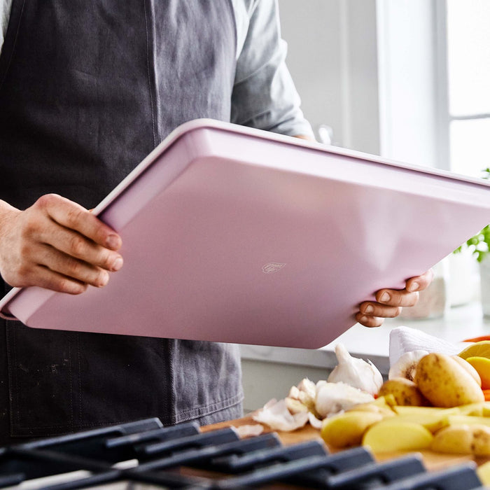 Person in an apron holds a pink ceramic 18 x 13 baking sheet above a kitchen counter.