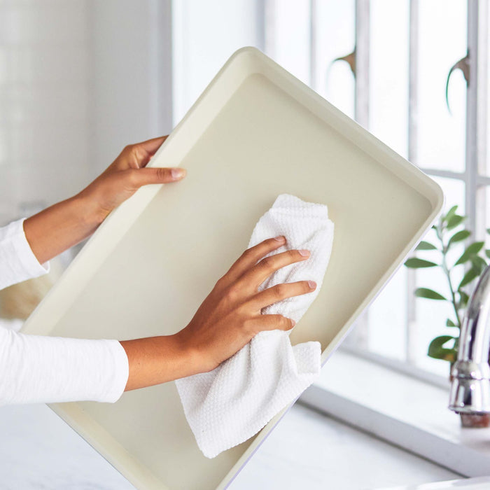 Pink ceramic nonstick cookie sheet being cleaned with a white cloth near a window and sink.